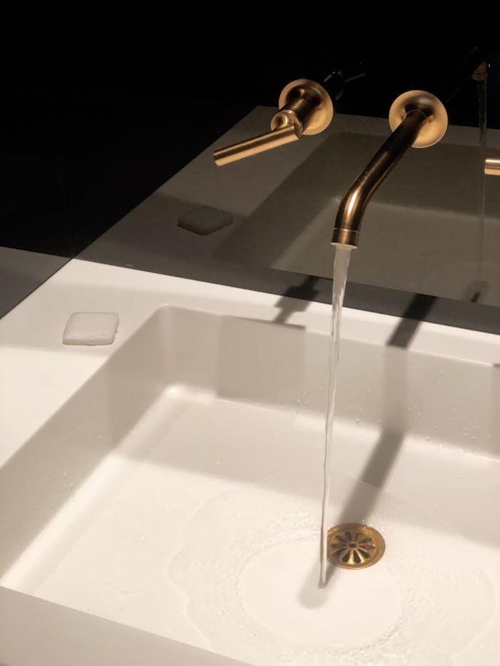 Home A sleek gold faucet pours water into a modern white washbasin in a dimly lit, elegant bathroom setting.